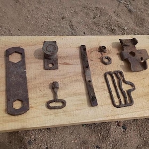 Puede incluir: Una colección de piezas de ferretería metálicas oxidadas, que incluyen una herramienta similar a una llave, un pequeño mango y varios soportes y sujetadores, dispuestas sobre una tabla de madera clara. Los artículos muestran signos de edad y desgaste.