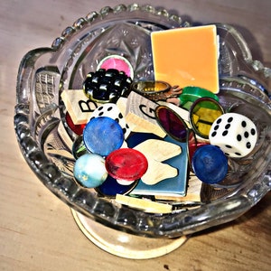 May include: A glass bowl filled with colorful game pieces, including dice, tiles, and counters. There is also a small orange square card in the bowl.