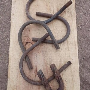 Puede incluir: Una colección de pernos en U y varillas curvas de metal oxidado se disponen sobre una tabla de madera de color claro. Las piezas metálicas tienen extremos roscados y muestran signos de edad y desgaste.