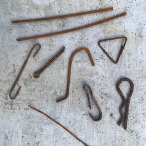 Puede incluir: Un surtido de piezas de ferretería metálicas oxidadas, incluyendo varillas, ganchos y un triángulo, se disponen sobre una superficie gris texturizada. Los elementos varían en tamaño y forma, mostrando un aspecto envejecido y desgastado.