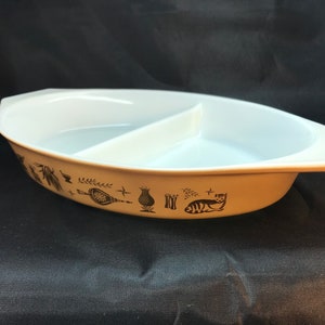 May include: Oval, divided serving dish with a cream-coloured exterior and white interior. The exterior features black decorative illustrations of kitchen items and a cat. The dish is designed for serving food.