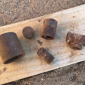 May include: A selection of rusty metal pipe fittings, including a coupling, a cap, a tee, and a nipple, arranged on a weathered wooden plank. The fittings show signs of age and corrosion, with a reddish-brown colour.