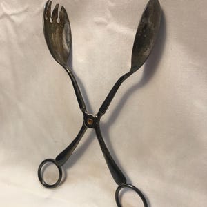 Puede incluir: Pinzas de servir plateadas con un extremo en forma de tenedor y otro en forma de cuchara, unidos en un pivote central. Los mangos tienen orificios circulares para los dedos. El metal muestra signos de uso.