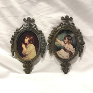 May include: Two ornate oval frames with gold accents, each featuring a vintage print of a young girl. One girl is praying, the other is holding a flower.