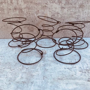 May include: A collection of six rusty metal springs, each with a circular base and a coiled, upright design. The springs are arranged on a textured, light-colored surface, creating an industrial or vintage aesthetic.