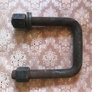 Peut inclure: Un grand boulon en U en métal gris foncé avec des extrémités filetées, doté de deux écrous hexagonaux et d'une rondelle plate fixée d'un côté. Cette fixation industrielle robuste présente une finition mate patinée et une base arrondie.
