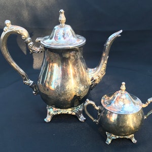 May include: A silver teapot and creamer set with ornate details and a polished finish. The teapot has a lid with a finial and a curved spout. The creamer has a lid and two handles.