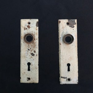 May include: Two antique white door plates with keyholes and round holes for door knobs. The plates are slightly worn and have a rustic look.