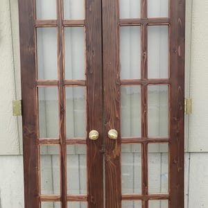 May include: A pair of dark brown stained wooden French doors featuring six glass panes per door. The doors are equipped with brass-colored metal hinges on the outer edges and two matching brass-colored round door knobs in the center.