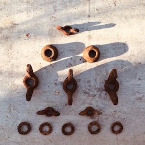 Puede incluir: Un surtido de piezas de ferretería metálica antiguas y desgastadas, que incluyen anillos, ojales y elementos decorativos, dispuestos sobre una superficie texturizada. Los artículos son de color marrón óxido, con signos visibles de edad y desgaste, lo que sugiere una estética vintage o de recuperación.