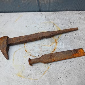 May include: Two rusty metal tools, a large spike and a smaller, flat tool, lie on a concrete surface. The spike has a broad head and a long, tapered body. The smaller tool is rectangular with a rounded end. Both show signs of age.