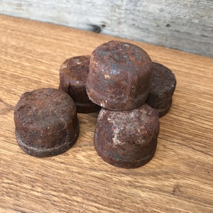 Op de afbeelding: Een verzameling van vijf kleine, cilindrische metalen pijpkappen met een zwaar verroest, donkerbruin en getextureerd oppervlak. Elke dop heeft een platte bovenkant en een licht verhoogde rand aan de basis, wat duidt op aanzienlijke oxidatie.