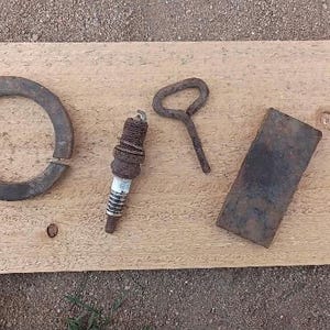 Op de afbeelding: Een verzameling vintage metalen voorwerpen gerangschikt op een lichtbruin houten bord. De items omvatten een roestige ring, een bougie, een flesopener en een rechthoekige metalen plaat. De objecten zijn verweerd en vertonen tekenen van ouderdom.