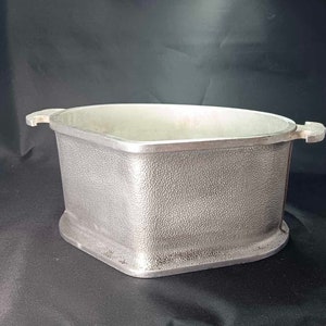 May include: A rectangular, silver-colored metal container with a textured surface. The container has a wide, open top and two small handles on either side. The base is slightly rounded.