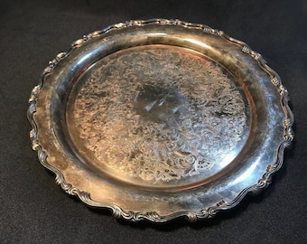 May include: Silver-toned metal serving tray with an ornate floral pattern in the center. The tray has a scalloped edge and a reflective surface. The tray is round and appears to be vintage.