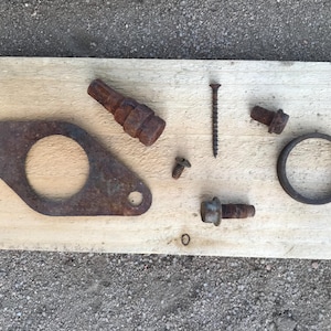 Peut inclure: Ensemble de pièces métalliques rouillées, comprenant des boulons, des vis, un joint et un anneau, disposées sur une planche de bois claire. Les articles sont de tailles et de formes variées, montrant des signes d'âge et d'usure.