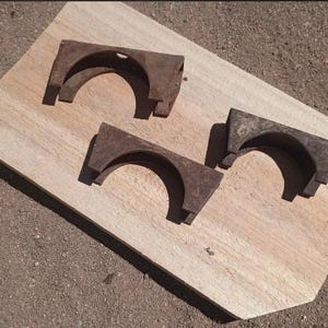 May include: Three identical, weathered, dark brown metal brackets with a semi-circular cutout in the center. Each bracket features a flat top edge and two downward-extending sides, showing signs of oxidation and a rough, aged texture throughout.