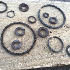Puede incluir: Una colección de varios anillos y arandelas metálicas oxidadas de diferentes tamaños, dispersos sobre una superficie de madera desgastada. Los elementos parecen viejos y desgastados, con signos de corrosión y envejecimiento. La disposición incluye formas circulares y semicirculares.