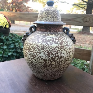 May include: A ceramic jar with a lid, featuring a crackled glaze in shades of brown and white. The jar has a rounded body and a decorative handle on each side.