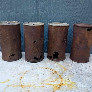 Może przedstawiać: Cztery zardzewiałe, cylindryczne metalowe puszki ze znacznymi śladami użytkowania i dziurami. Puszki są brązowe ze srebrnymi obwódkami. Stoją na betonowej powierzchni na tle niebieskiego drewnianego tła.
