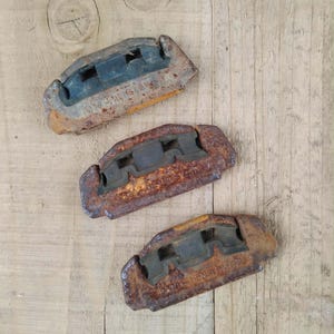 Könnte beinhalten: Drei rechteckige Metallbeschläge mit starker Rostoxidation und dunkelgrauen Gummieinsätzen. Jedes Teil weist einen mittigen Ausschnitt und geprägten Text entlang der Unterkante auf und zeigt deutliche Gebrauchs- und Altersspuren.