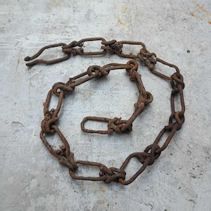 May include: A rusty metal chain with oval links, arranged in a spiral on a concrete surface. The chain shows signs of age and wear, with visible rust and discoloration. The links are connected with knots.