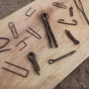 May include: A collection of various rusty metal fasteners, including cotter pins, paper clips, and staples, arranged on a light-colored wooden board. The fasteners are in different shapes and sizes, showing signs of age and wear.
