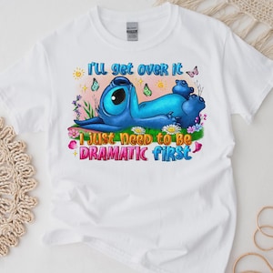 Peut inclure: T-shirt blanc avec un personnage de dessin animé bleu allongé sur de l'herbe avec des fleurs et des papillons. Le texte sur le t-shirt dit : "Je vais surmonter ça, j'ai juste besoin d'être dramatique d'abord."