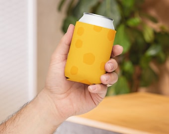 May include: A yellow drinks can cooler with a cheese pattern is held in a hand. The cooler is designed to fit a standard-sized can. The background includes a plant and a wooden surface.