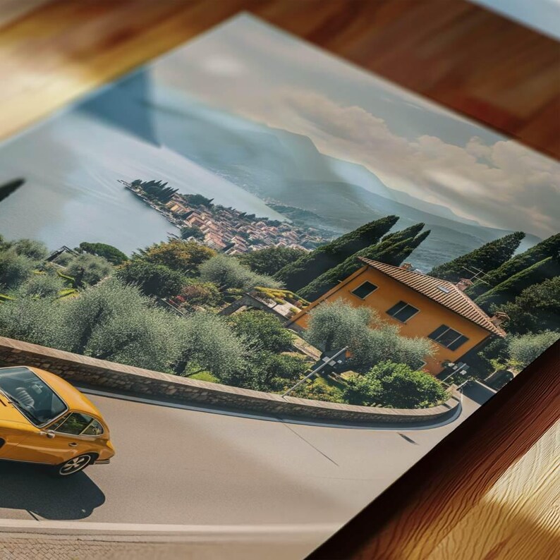 May include: A print depicting a coastal scene with a yellow classic car on a winding road. The image shows a town by a lake, with green trees, an orange building, and mountains in the background, creating a scenic landscape.