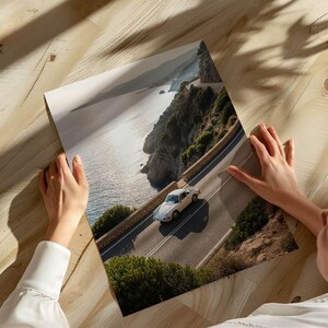 May include: A print depicting a white classic car on a coastal road. The image shows a scenic view of the sea, cliffs, and road. The car is the central element, set against a backdrop of natural beauty.