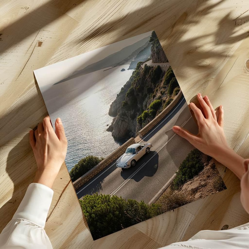 May include: A print depicting a white classic car on a coastal road. The image shows a scenic view of the sea, cliffs, and road. The car is the central element, set against a backdrop of natural beauty.