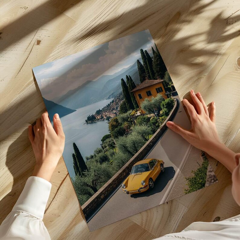 May include: A scenic print depicting a yellow sports car on a winding road, with a lake and mountains in the background. The print shows an Italian landscape with a villa and tall trees. The print is held by a person.