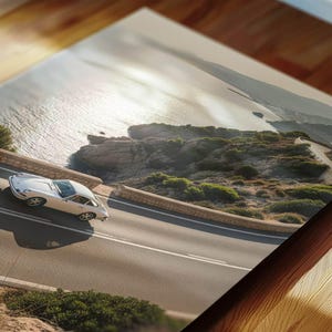 May include: A white vintage sports car on a coastal road, with the sea and cliffs in the background. The image is a print on a wooden surface, showcasing the car's design and the scenic route. The road curves along the coastline.