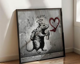 May include: Framed artwork depicting a black and white illustration of a rat with a backpack and halo, spraying a red heart on a distressed wall. The artwork is in a black frame, leaning against a white wall and wooden floor.