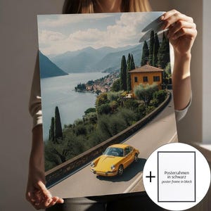 May include: A poster print depicting a yellow vintage car on a road beside a lake, with a yellow building and tall trees. The background shows a town on the water's edge and a blue sky with clouds. The print includes a black poster frame.