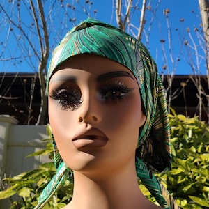 May include: A green and gold patterned satin head wrap with a gathered knot on top. The head wrap is worn on a mannequin head.