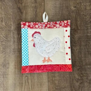 May include: Square quilted potholder featuring a white chicken applique with red comb and feet. The potholder has a cream-colored center, bordered by red floral and polka dot fabrics, and a blue polka dot stripe. A white loop is attached for hanging.