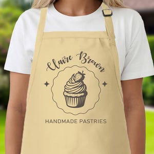 May include: A beige apron featuring the name "Claire Brown" and the words "Handmade Pastries" in black script. A cupcake illustration with a strawberry is in the center. The apron has a pocket and an adjustable neck strap.