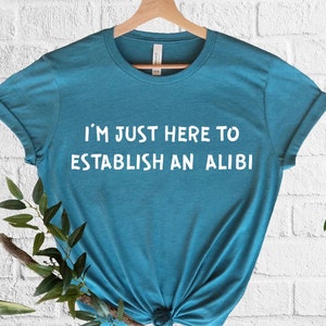 May include: A teal blue t-shirt with the text "I'm just here to establish an alibi" printed in white on the front.