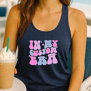 May include: Navy blue tank top with the text "IN MY CUSTOM ERA" in pink and blue. The woman in the image is holding a blended coffee drink with a green straw. The background is a beach scene.
