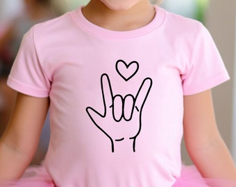May include: A young girl wearing a pink tutu and a pink shirt with a black hand sign for 'I love you' and a small heart above it. She is wearing a sparkly gold crown.