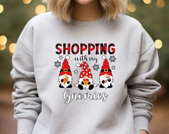 May include: A light gray crewneck sweatshirt featuring a graphic print of three gnomes wearing red hats. The text reads SHOPPING with my Gnomies in black and red plaid lettering. The gnomes hold a gingerbread cookie, a gift, and a candy cane.