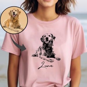 May include: A pink t-shirt featuring a black and white illustration of a dog. The dog is lying down and the text "Luna" is printed below the dog.
