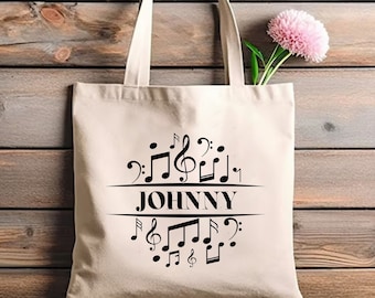 May include: A beige tote bag with black musical notes and the name "JOHNNY" printed in the center. A pink flower is placed inside the bag. The bag has two white handles and is set against a wooden background.