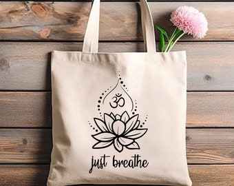May include: Cream-colored tote bag with a black lotus flower design and the words "just breathe." The bag has a simple handle and is made of a sturdy fabric. A pink flower is placed inside the bag.