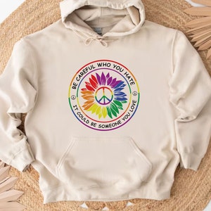 Pu&ograve; includere: Una felpa con cappuccio beige con una grafica di girasole arcobaleno e un simbolo di pace. La grafica include il testo "Fai attenzione a chi odi, potrebbe essere qualcuno che ami".