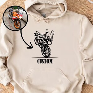 May include: A cream-colored hoodie with a black illustration of a motorcycle and the word "CUSTOM" printed on the front.