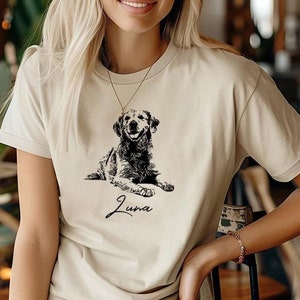 May include: A beige t-shirt featuring a black and white illustration of a dog sitting down. The dog's name, "Luna", is written in cursive below the illustration.