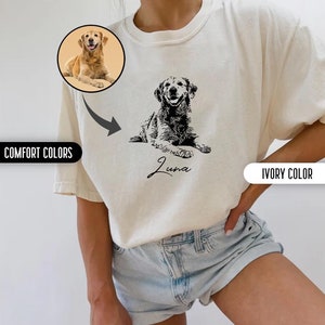 K&ouml;nnte beinhalten: Ein wei&szlig;es T-Shirt mit einer schwarz-wei&szlig;en Illustration eines Hundes und dem Namen "Luna" darauf gedruckt. Das Shirt wird von einer Person in Jeans-Shorts getragen.